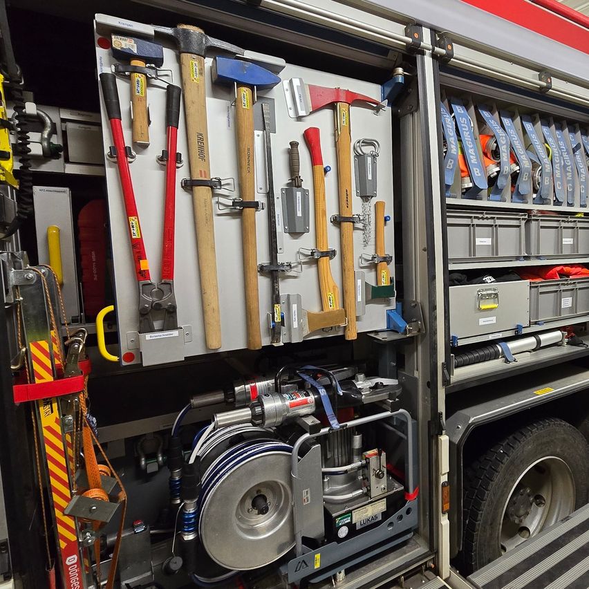 Der offene Bereich eines Feuerwehrwagens zeigt verschiedene Werkzeuge wie Hämmer, Äxte und ein großes silbernes Rad, zusammen mit Aufbewahrungsboxen und Sicherheitsausrüstung.