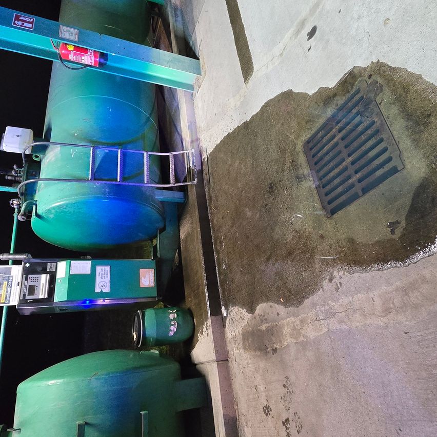 Eine Nahaufnahme einer Betonwand mit einer Wasserpfütze. Links ist eine grüne Maschine mit einer Leiter und einem roten Feuerlöscher angebracht.