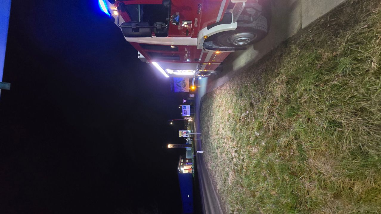 Ein Feuerwehrauto ist nachts am Straßenrand geparkt, mit eingeschalteten Lichtern. Das Auto ist rot mit weißen Streifen und steht auf einer abschüssigen Oberfläche.