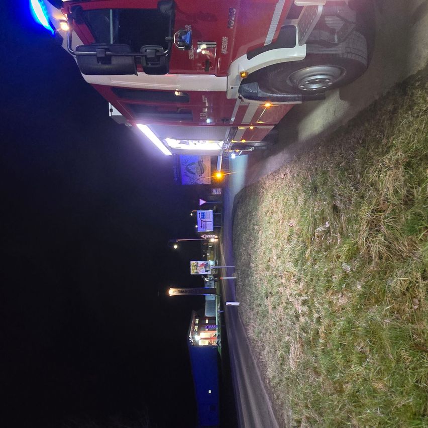 Ein Feuerwehrauto ist nachts am Straßenrand geparkt, mit eingeschalteten Lichtern. Das Auto ist rot mit weißen Streifen und steht auf einer abschüssigen Oberfläche.