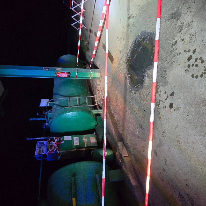 Ein nächtlicher Blick auf einen Lagerbereich mit grünen Tanks, einem Feuerlöscher und einem Loch in der Wand. Rote und weiße Stangen sind in der Nähe positioniert.