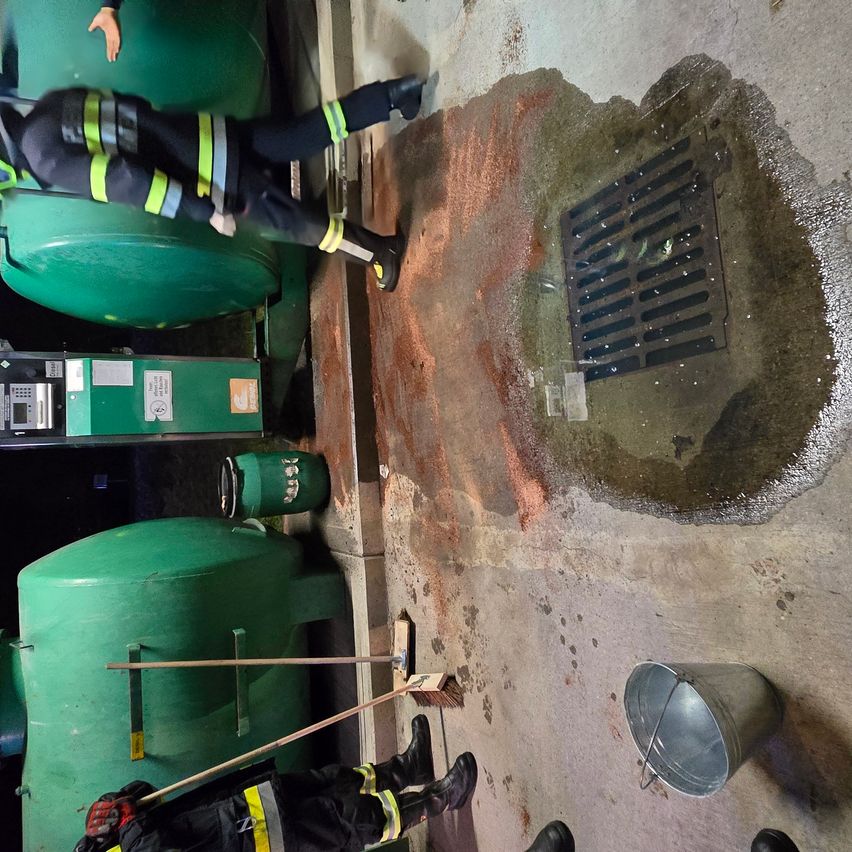 Ein Feuerwehrmann reinigt einen Ausguss auf dem Boden in der Nähe eines Wassertanks. Ein Eimer und ein Besen sind in der Nähe.