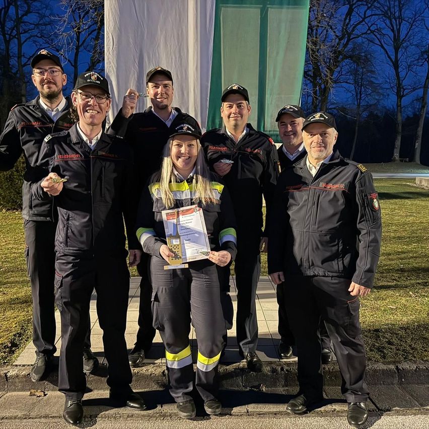 Eine Gruppe von Feuerwehrleuten posiert für ein Foto im Freien. Sie tragen alle Uniformen. Eine Frau in der Mitte hält ein Zertifikat.
