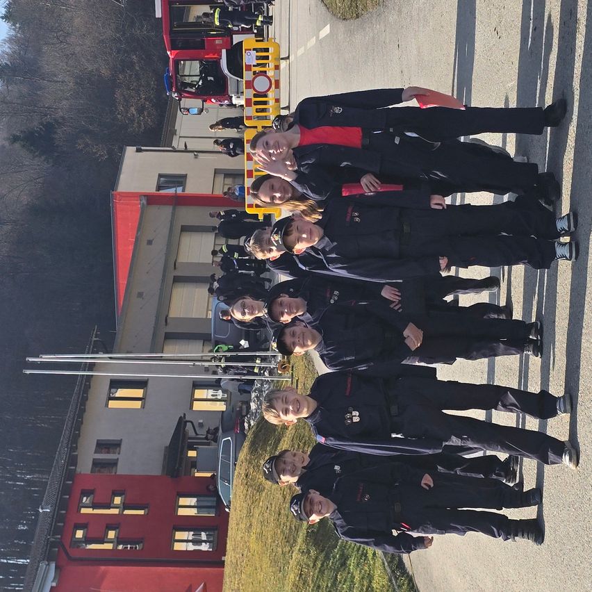 Eine Gruppe von Menschen in Uniformen steht in einer Reihe vor einer Feuerwache. Sie lächeln und posieren für ein Foto.