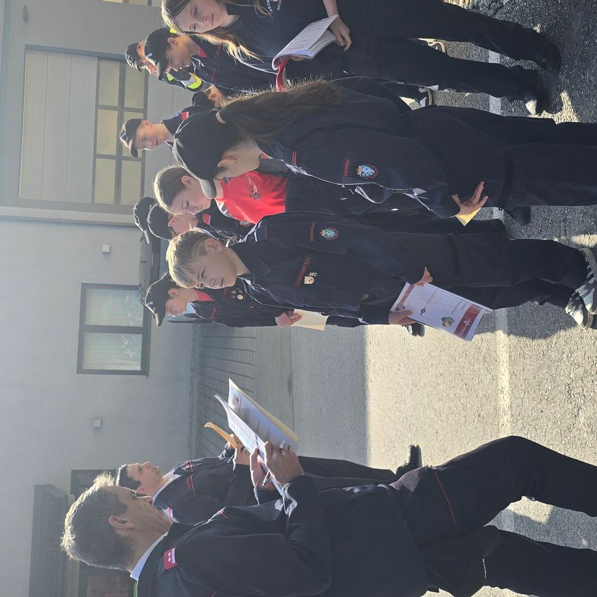 Eine Gruppe von Personen in Uniformen, möglicherweise für eine Zeremonie, steht in einer Reihe vor einem Gebäude. Sie halten Papiere in der Hand und scheinen jemandem zuzuhören.