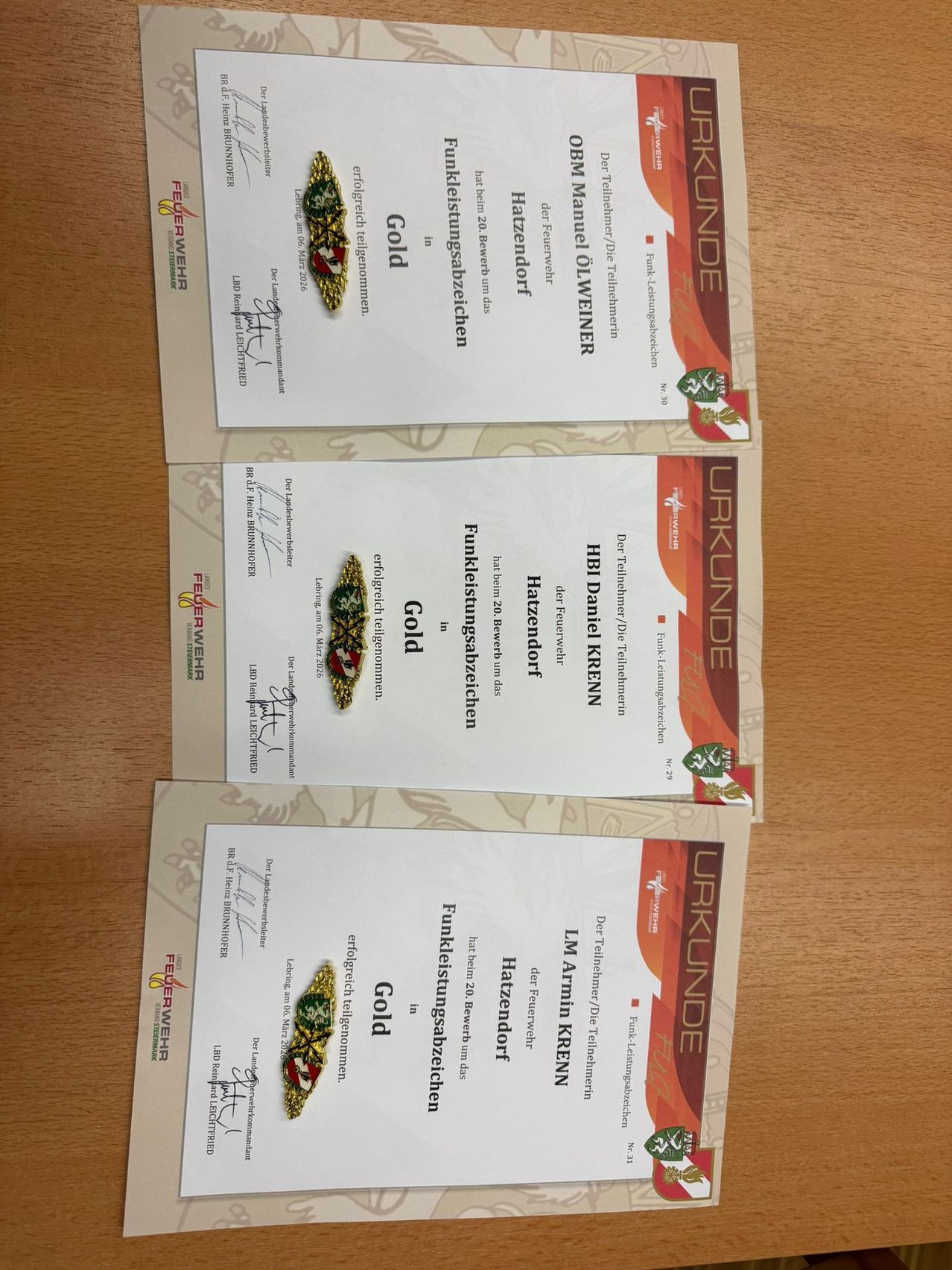 Drei Zertifikate liegen auf einer Holzoberfläche. Jedes Zertifikat hat ein goldenes Emblem und ist mit den Wörtern 'Feuerwehr' und 'Gold' beschriftet. Der Hintergrund der Zertifikate ist weiß mit roten und schwarzen Rändern.