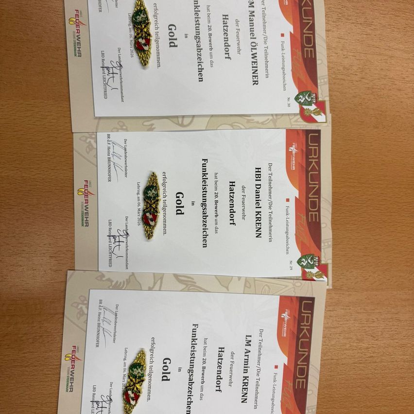 Drei Zertifikate liegen auf einer Holzoberfläche. Jedes Zertifikat hat ein goldenes Emblem und ist mit den Wörtern 'Feuerwehr' und 'Gold' beschriftet. Der Hintergrund der Zertifikate ist weiß mit roten und schwarzen Rändern.