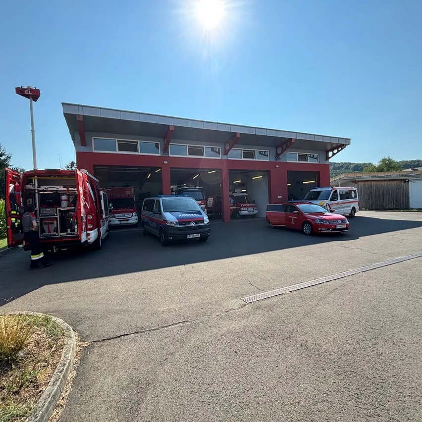 Eine Feuerwehrstation mit mehreren Rettungsfahrzeugen, die unter der hellen Sonne draußen geparkt sind. Eine Person steht in der Nähe eines Feuerwehrwagens.