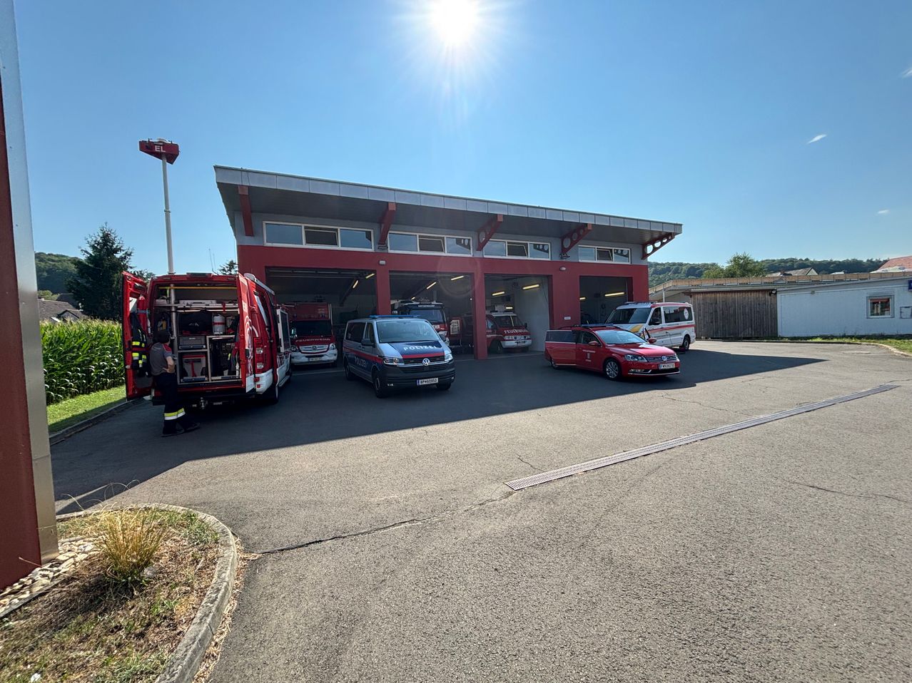 Eine Feuerwehrstation mit mehreren Rettungsfahrzeugen, die unter der hellen Sonne draußen geparkt sind. Eine Person steht in der Nähe eines Feuerwehrwagens.