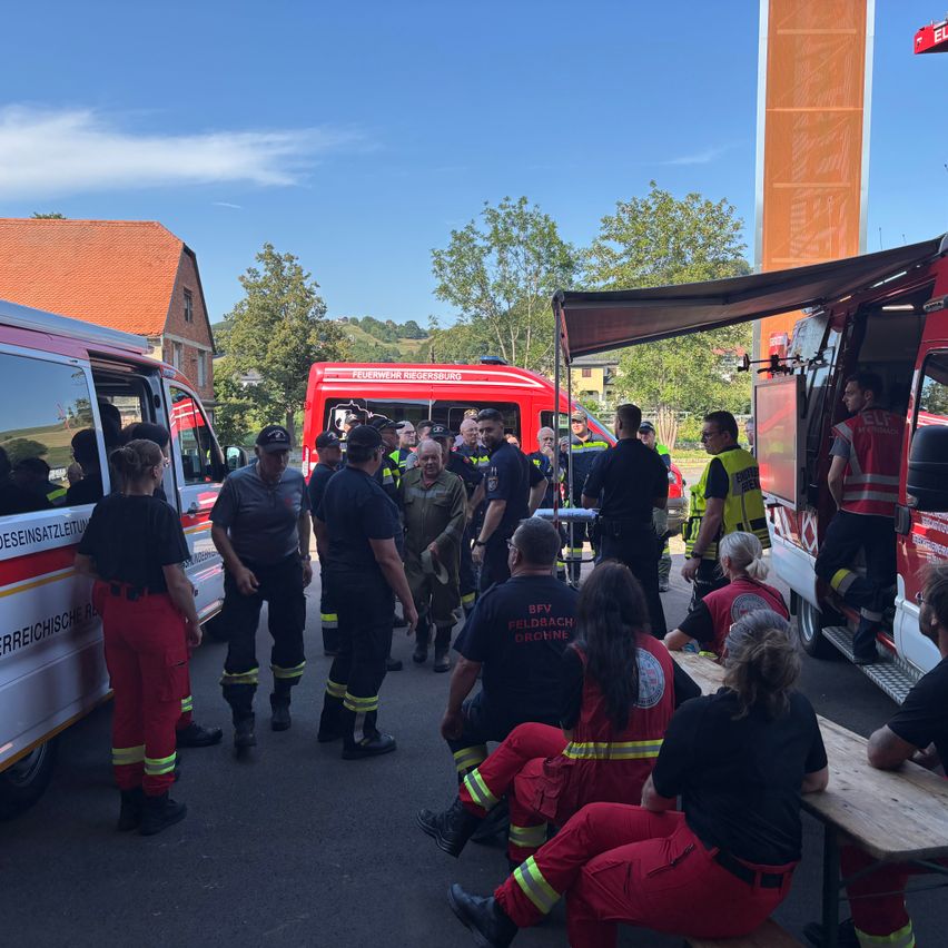 Eine Gruppe von Feuerwehrleuten und Rettungskräften versammelt sich um einen roten Feuerwehrwagen und einen roten Krankenwagen. Einige sitzen auf Bänken, andere stehen. Bäume und ein Gebäude sind im Hintergrund.