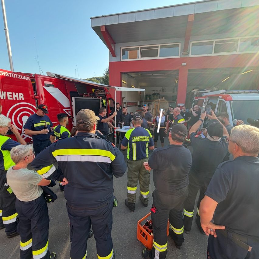 Eine Gruppe von Feuerwehrleuten und Zivilisten versammelt sich vor einer Feuerwache, einige halten Getränke und andere machen Fotos.