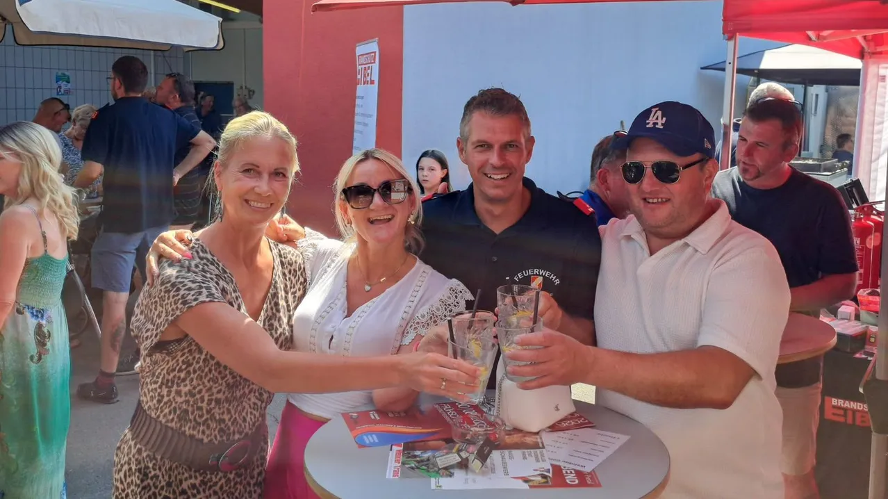 Vier Erwachsene, zwei Frauen und zwei Männer, lächeln und halten Getränke an einem Tisch, mit einem Banner und Menschen im Hintergrund.