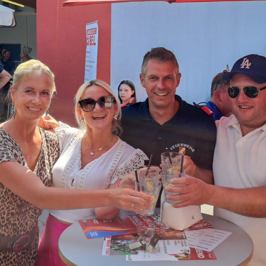 Vier Erwachsene, zwei Frauen und zwei Männer, lächeln und halten Getränke an einem Tisch, mit einem Banner und Menschen im Hintergrund.