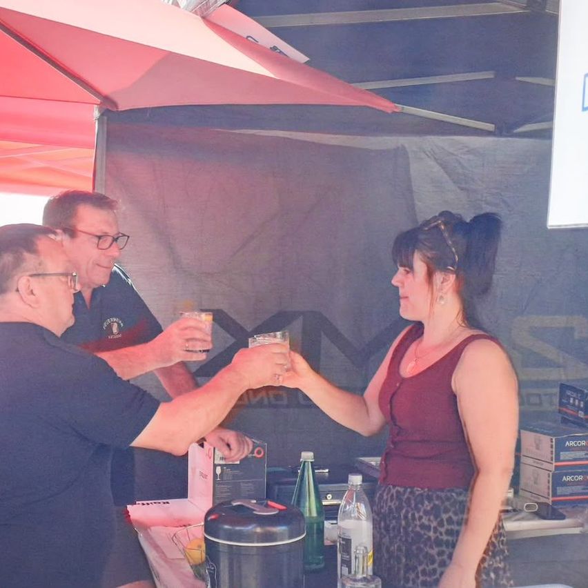 Drei Personen stehen unter einem Zelt. Zwei Männer halten Gläser mit Getränken und eine Frau hält auch ein Glas. Sie lächeln und scheinen Spaß zu haben. Auf dem Tisch stehen Flaschen und Kisten.