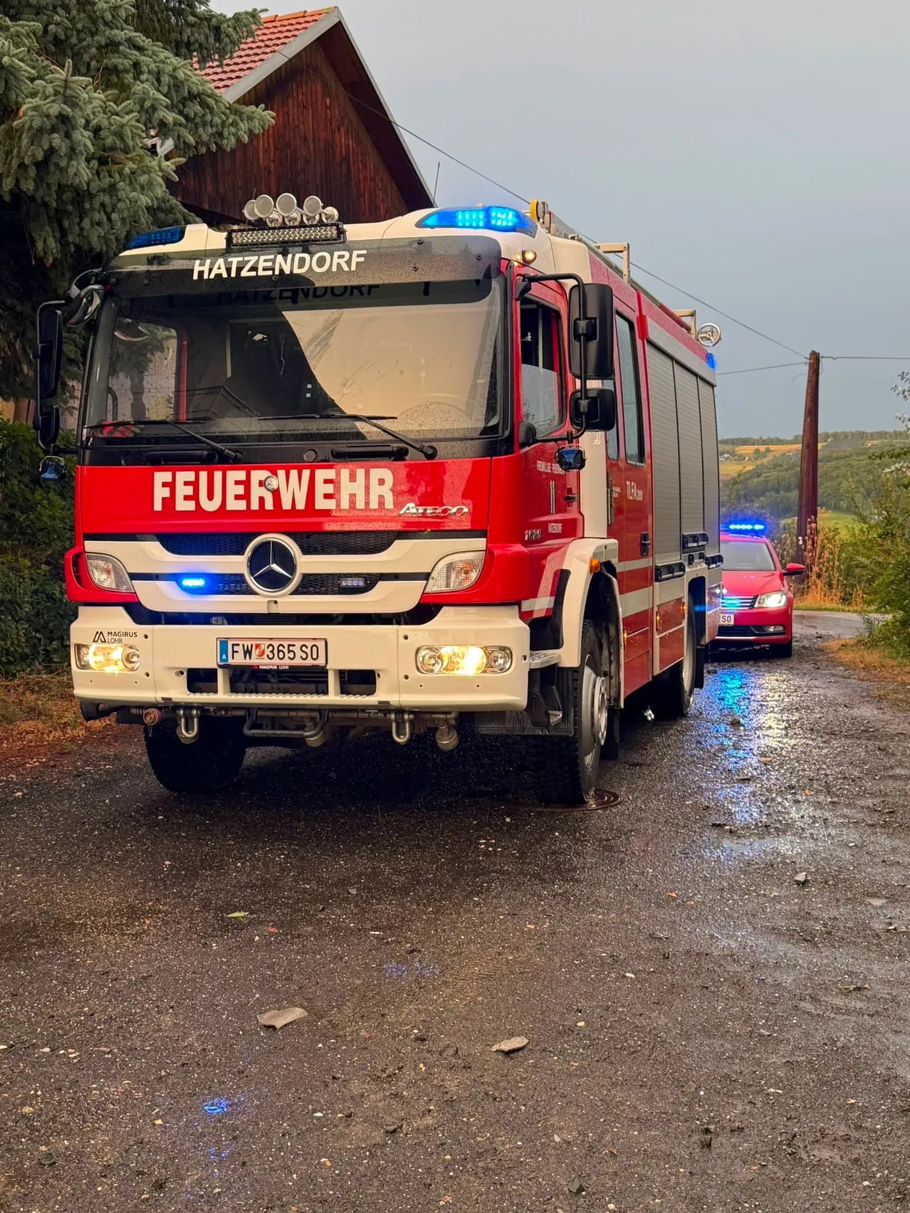 Ein roter Feuerwehrwagen aus Hatzendorf mit dem Wort 'Feuerwehr' auf der Front ist auf einer nassen Straße geparkt. Ein rotes Auto ist dahinter geparkt.