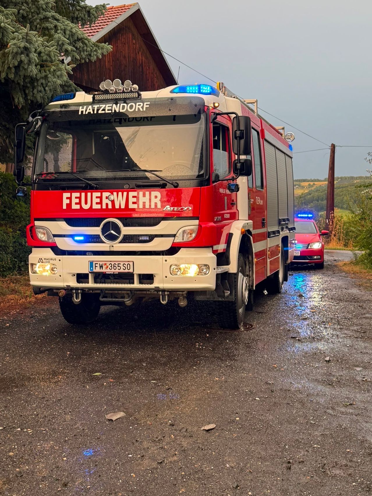 Ein roter Feuerwehrwagen aus Hatzendorf mit dem Wort 'Feuerwehr' auf der Front ist auf einer nassen Straße geparkt. Ein rotes Auto ist dahinter geparkt.