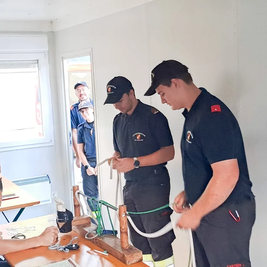 Drei Feuerwehrleute üben das Knotenlegen in einem Schulungsraum mit einem Tisch und einem Fenster im Hintergrund.