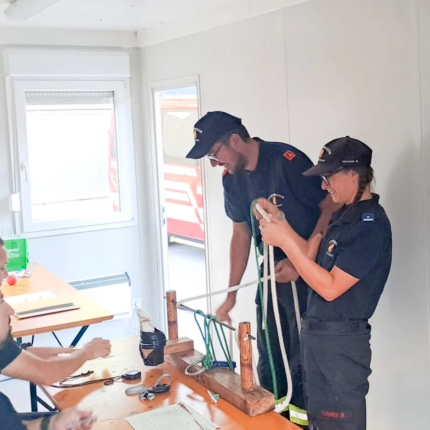 Zwei Feuerwehrleute in Uniform üben das Knotenlegen an einer Holzplatte in einem Raum. Eine andere Person sitzt an einem Schreibtisch mit Papieren und einem roten Apfel.