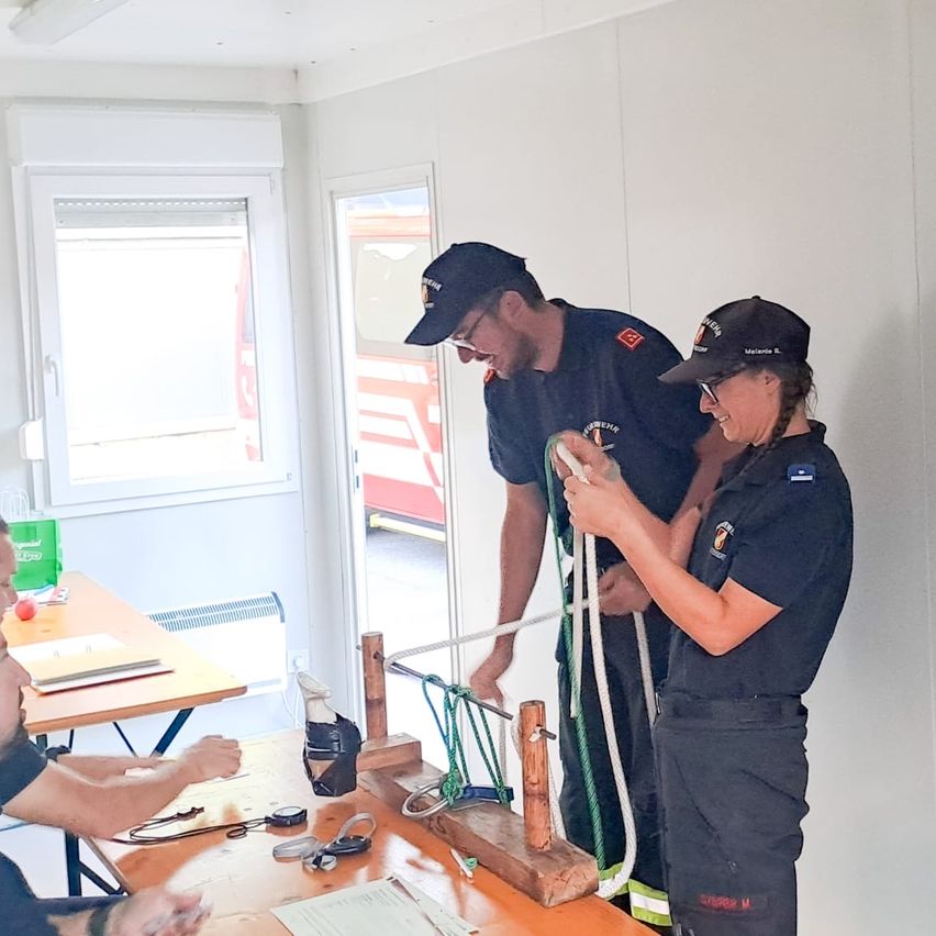Zwei Feuerwehrleute in Uniform üben das Knotenlegen an einer Holzplatte in einem Raum. Eine andere Person sitzt an einem Schreibtisch mit Papieren und einem roten Apfel.