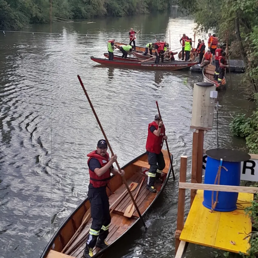 Eine Gruppe von Menschen in roten Rettungswesten und Mützen rudert auf einem Fluss. Ein Mann steht im vorderen Boot und hält eine Paddel. Andere Boote sind in der Nähe angedockt.