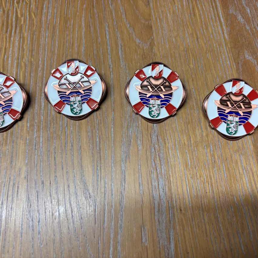Fünf runde Abzeichen mit rot-weißen karierten Rändern, jedes mit einem zentralen Emblem eines Bootes und Ruder, auf einer Holzoberfläche ausgestellt.