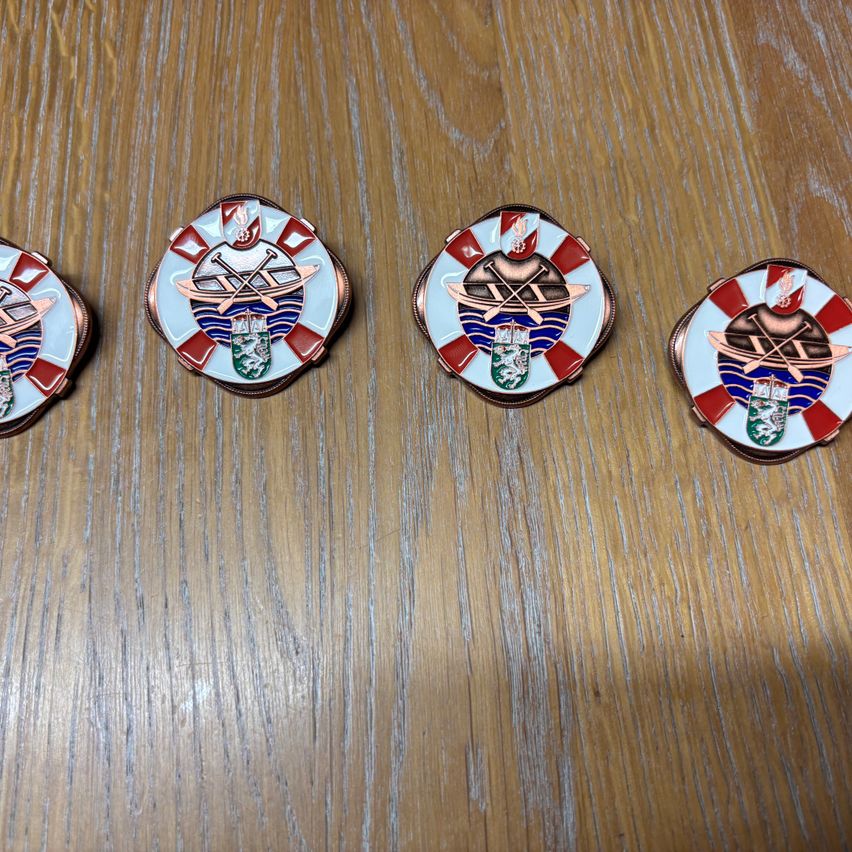 Fünf runde Abzeichen mit rot-weißen karierten Rändern, jedes mit einem zentralen Emblem eines Bootes und Ruder, auf einer Holzoberfläche ausgestellt.