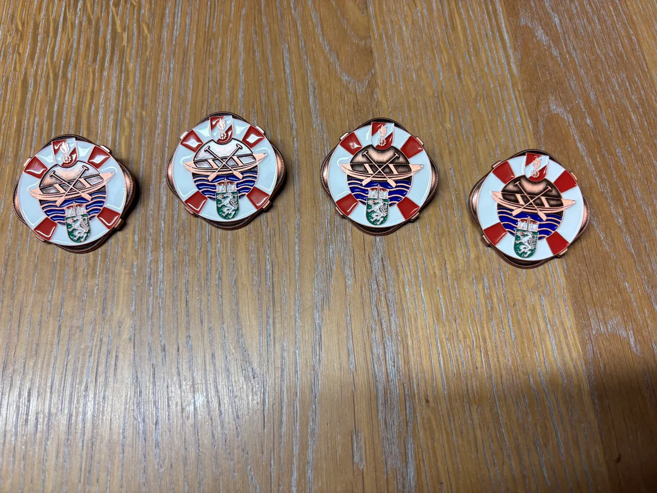 Fünf runde Abzeichen mit rot-weißen karierten Rändern, jedes mit einem zentralen Emblem eines Bootes und Ruder, auf einer Holzoberfläche ausgestellt.