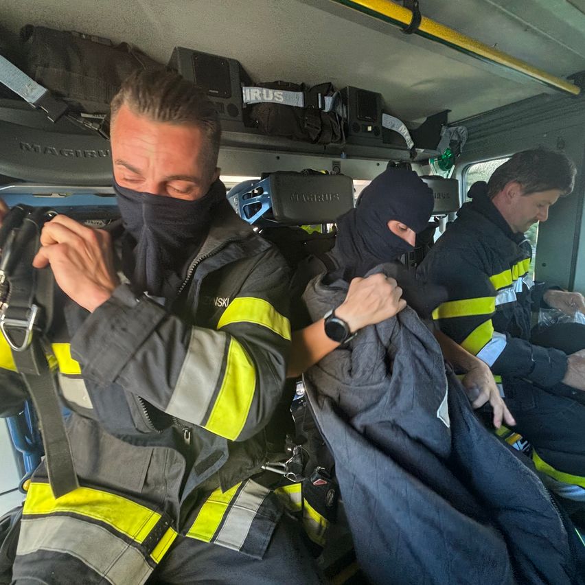 Drei Feuerwehrleute in Schutzkleidung in einem Fahrzeug, einer justiert seine Maske, während ein anderer die Jacke einer Person hält.