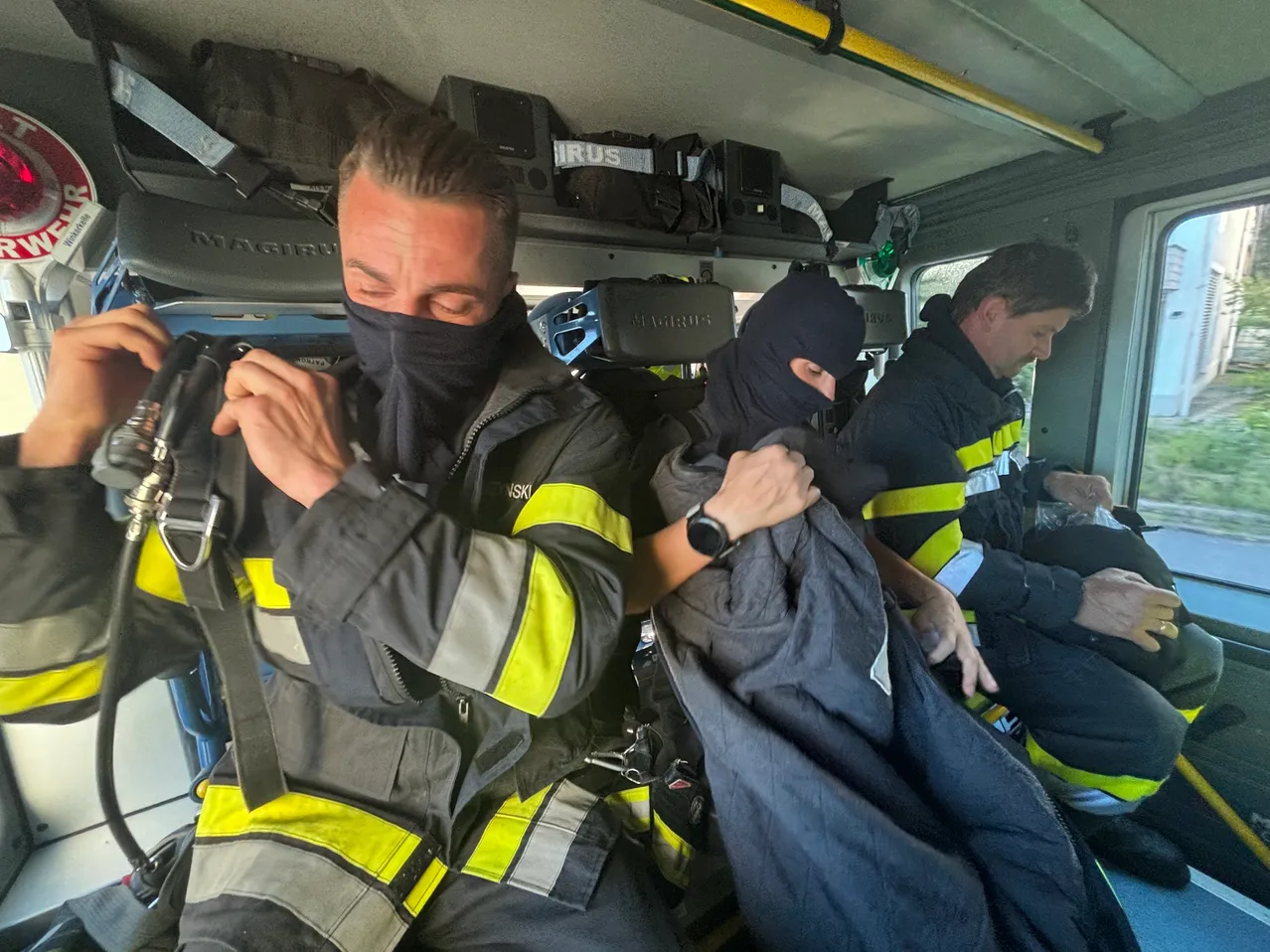 Drei Feuerwehrleute in Schutzkleidung in einem Fahrzeug, einer justiert seine Maske, während ein anderer die Jacke einer Person hält.