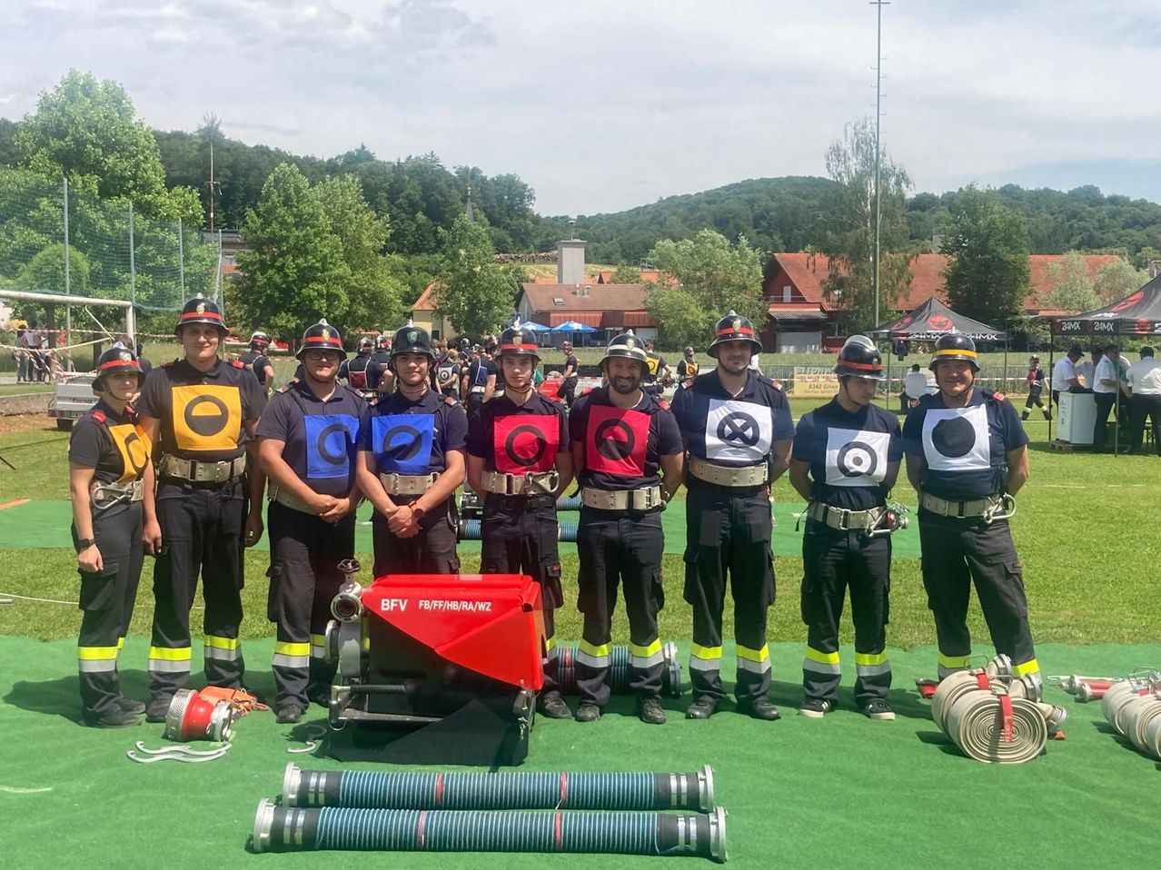 Eine Gruppe Feuerwehrleute steht auf einem grünen Feld, trägt Helme und Uniformen mit Sicherheitswesten. Sie halten eine rote Maschine mit dem Text BFV. Im Hintergrund befinden sich viele Bäume und Häuser.