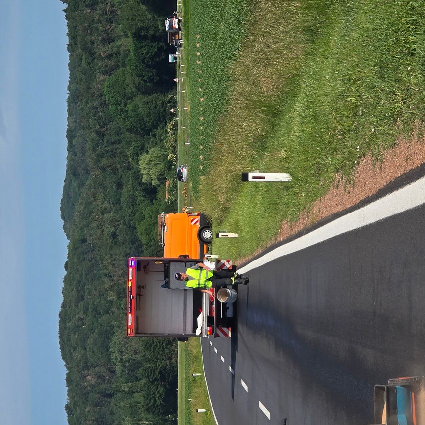 Luftaufnahme von Arbeitern in Sicherheitskleidung, die neue Asphalt auf einer kurvigen Straße auftragen, mit einer hügeligen Landschaft im Hintergrund.