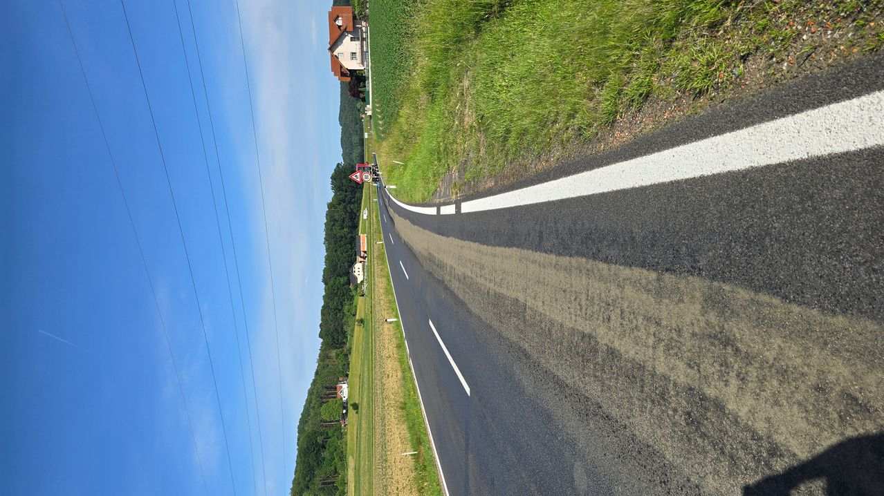 Luftaufnahme einer Landstraße mit weißen Markierungen und einem Haus in der Ferne. Die Straße wird von grasbewachsenen Feldern und Bäumen gesäumt.