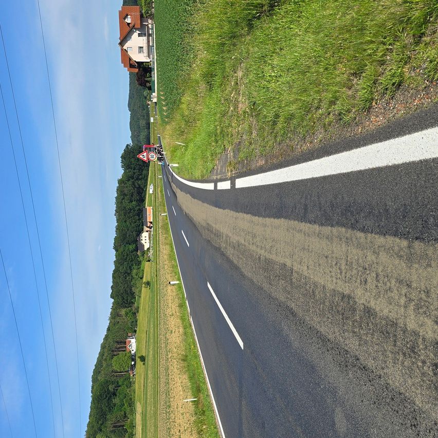 Luftaufnahme einer Landstraße mit weißen Markierungen und einem Haus in der Ferne. Die Straße wird von grasbewachsenen Feldern und Bäumen gesäumt.