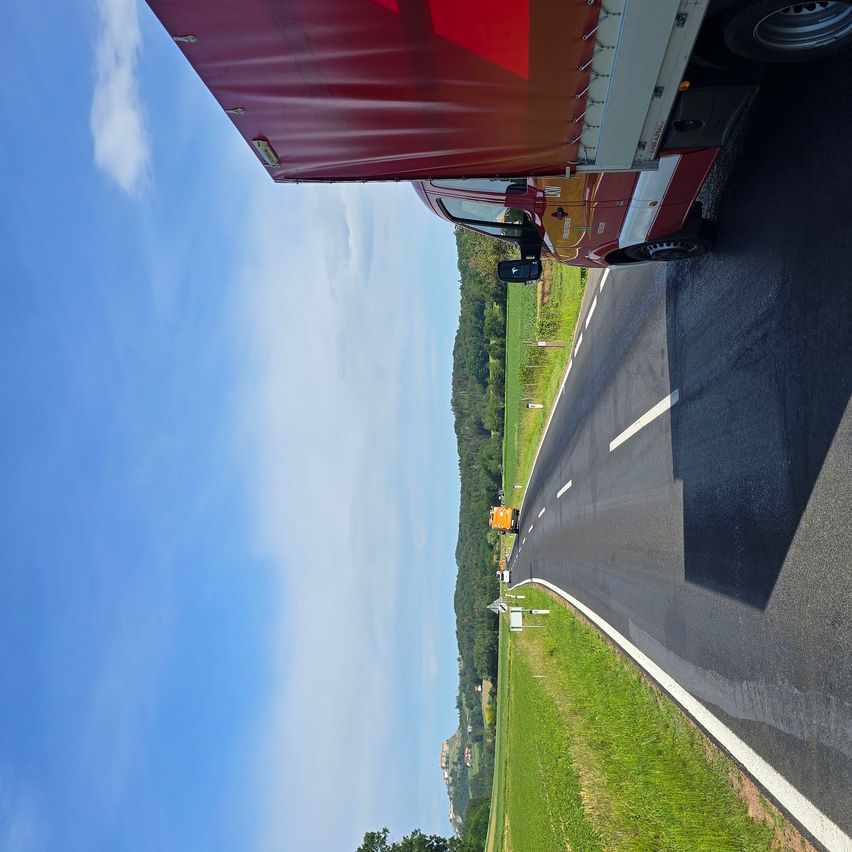 Ein Lkw mit einem roten Anhänger ist auf einer Straße, mit klarem blauem Himmel und Wolken darüber. Ein Fahrzeug ist in der Ferne auf der linken Seite.