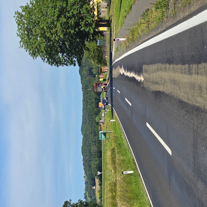 Eine Luftaufnahme einer im Bau befindlichen Straße mit Arbeitern in Sicherheitswesten. Ein Baufahrzeug ist am Straßenrand geparkt. Bäume und Gras säumen die Straße.