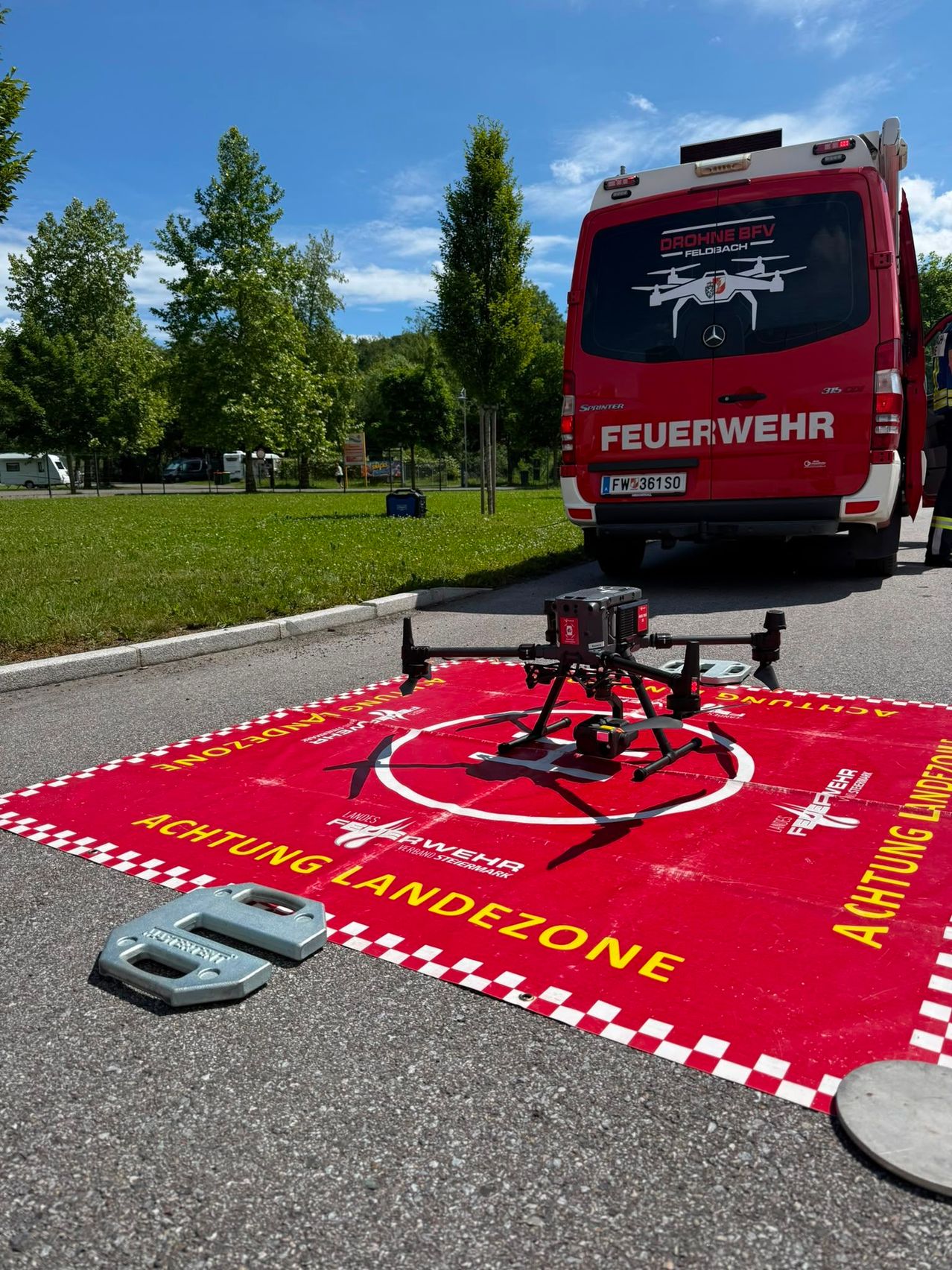 Eine rote Landezone für Drohnen ist auf dem Boden. Ein Feuerwehrwagen ist hinter der Landezone geparkt. Bäume und Gras sind im Hintergrund.