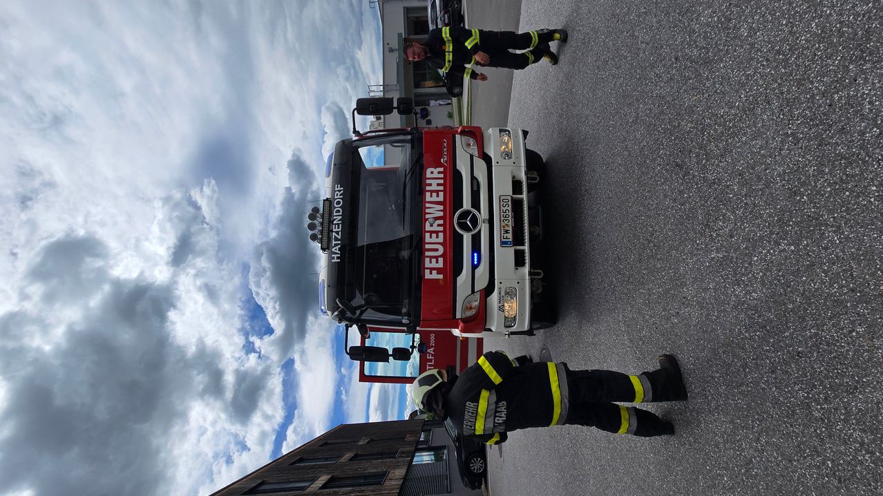Ein Feuerwehrwagen mit der Aufschrift Hatzendorf ist auf der Straße geparkt. Zwei Feuerwehrleute stehen in der Nähe des Fahrzeugs, einer hält einen Schlauch. Der Himmel ist bewölkt.