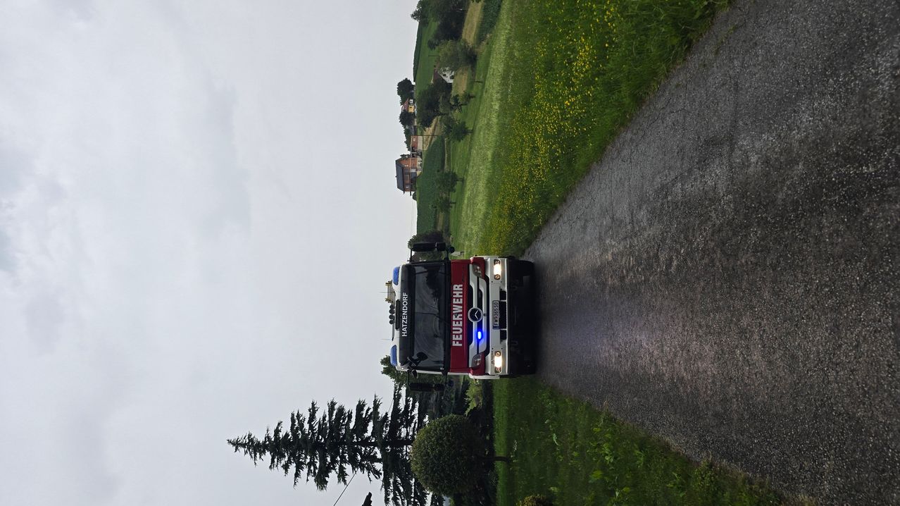 Ein Feuerwehrfahrzeug steht am Straßenrand, mit grünen Feldern und Häusern im Hintergrund. Das Fahrzeug hat das Wort Feuerwehr an der Seite.