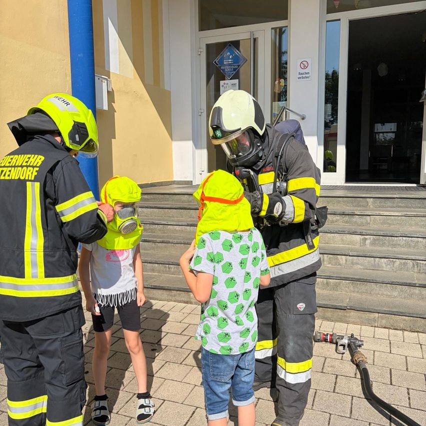Feuerwehrleute helfen Kindern beim Anprobieren von Gasmasken. Ein Kind trägt eine Maske auf dem Kopf, ein anderes auf dem Gesicht. Sie stehen vor einem Gebäude.