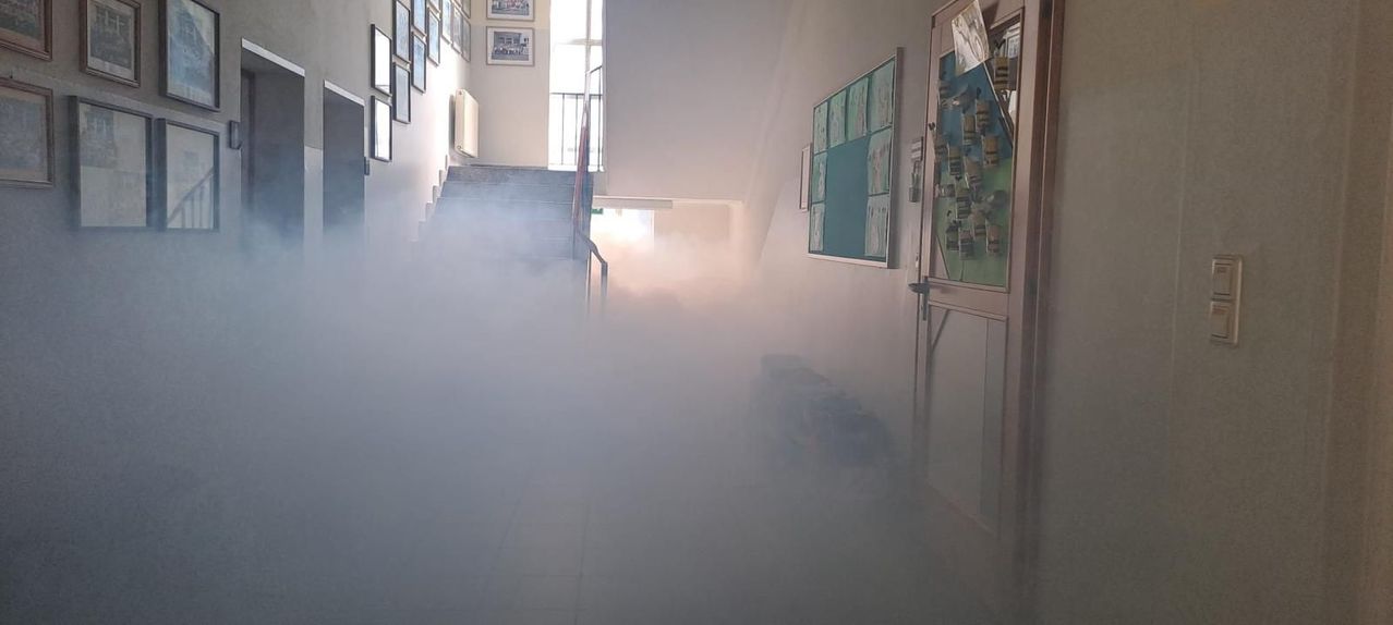 Ein Flur gefüllt mit dichtem Nebel. Treppen und ein Fenster sind sichtbar. Gerahmte Bilder schmücken die Wand, und eine Pinnwand befindet sich rechts.