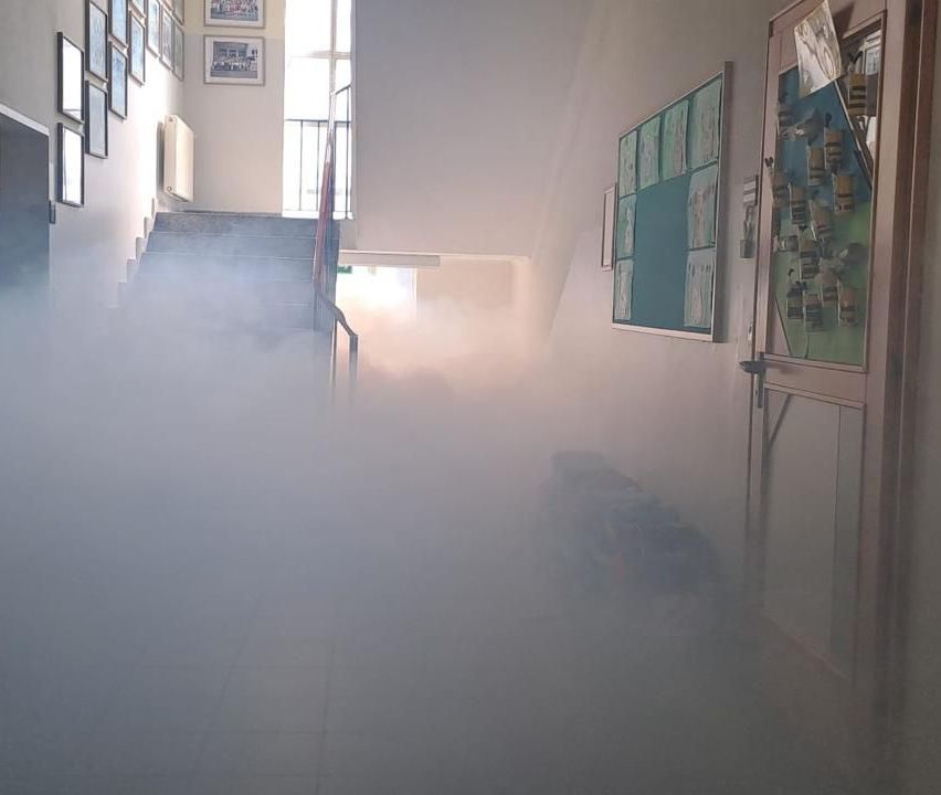 Ein Flur gefüllt mit dichtem Nebel. Treppen und ein Fenster sind sichtbar. Gerahmte Bilder schmücken die Wand, und eine Pinnwand befindet sich rechts.
