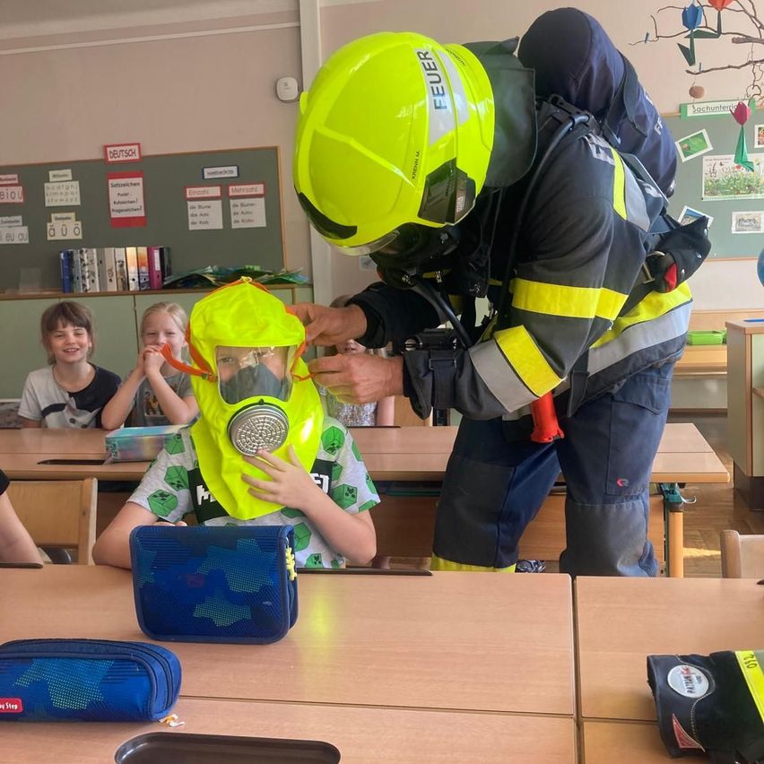 Ein Feuerwehrmann in voller Ausrüstung justiert eine Gasmaske eines Kindes in einem Klassenzimmer, während andere Kinder zusehen.