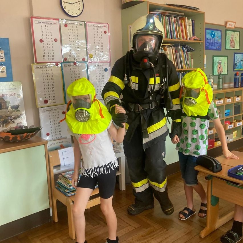 Ein Feuerwehrmann in voller Ausrüstung steht mit zwei Kindern in einem Klassenzimmer. Beide Kinder tragen neongelbe Gasmasken und der Feuerwehrmann hat ebenfalls eine Maske auf.