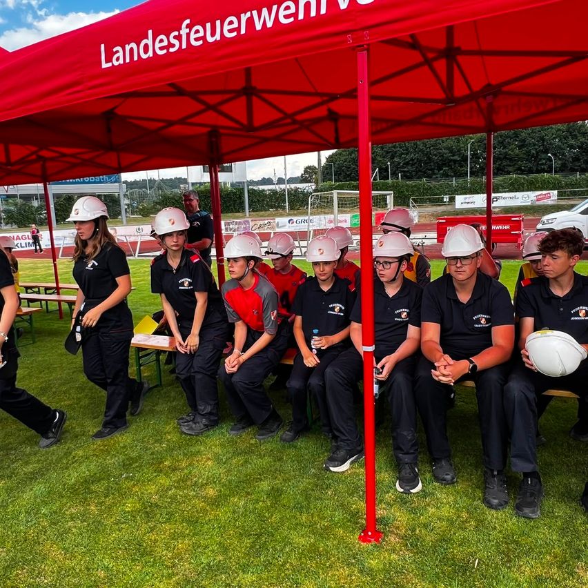 Eine Gruppe junger Menschen in Uniformen und Helmen sitzt unter einem roten Zelt auf einem Rasenfeld.
