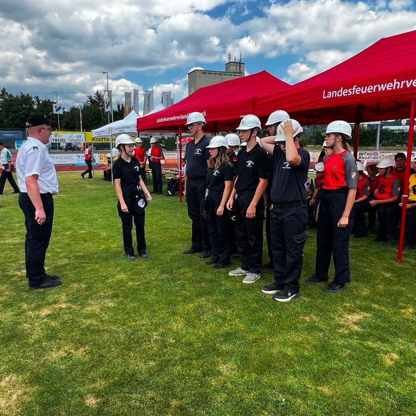 Eine Gruppe von Menschen in Uniform, möglicherweise Feuerwehrleute, stehen in einer Reihe unter einem roten Zelt auf einem Rasenfeld.