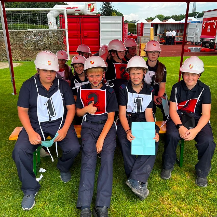 Eine Gruppe von Kindern in Helmen und Uniformen sitzt auf einer Bank, möglicherweise für ein Foto. Sie halten Papiere und lächeln. Dahinter steht eine rote Struktur mit einem Netz.