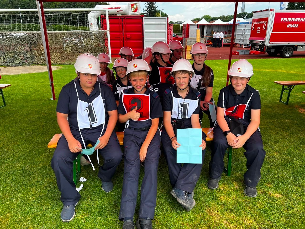 Eine Gruppe von Kindern in Helmen und Uniformen sitzt auf einer Bank, möglicherweise für ein Foto. Sie halten Papiere und lächeln. Dahinter steht eine rote Struktur mit einem Netz.