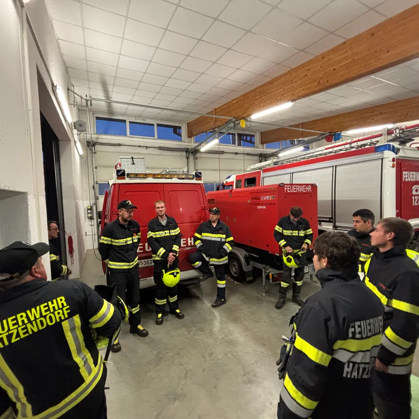 Eine Gruppe von Feuerwehrleuten in Uniform steht in einer Feuerwache, mit Feuerwehrautos im Hintergrund.