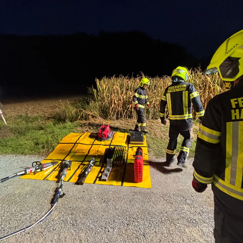 Drei Feuerwehrleute in gelben Helmen und reflektierenden Uniformen stehen vor einem Maisfeld bei Nacht. Sie bereiten Ausrüstung auf einem gelben Teppich auf dem Boden vor.