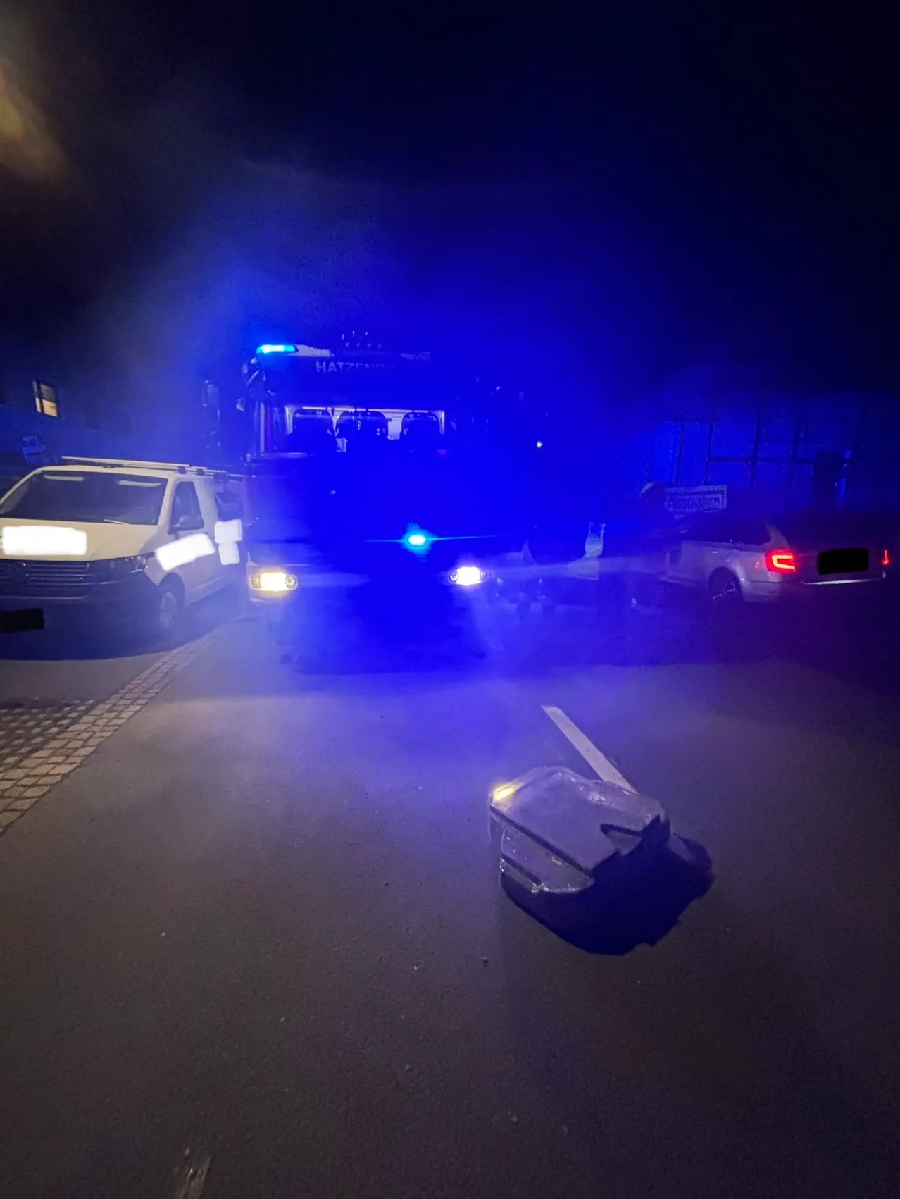 Ein Polizeibus mit blauen Lichtern ist am Straßenrand geparkt. Ein weißer Lieferwagen und ein Auto sind in der Nähe geparkt. Ein Beutel liegt auf der Straße.